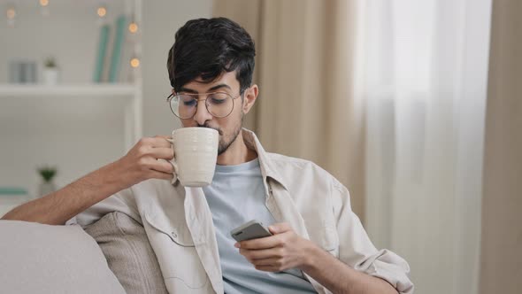 Young Relaxed Guy Indian Arabic Bearded Handsome Man Sitting at Home on Couch Drinking Tea Coffee alt