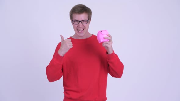 Happy Young Handsome Man Holding Piggy Bank and Giving Thumbs Up alt