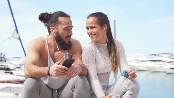 Couple Listening To Music Together Using One Earphones Sitting on Pier alt