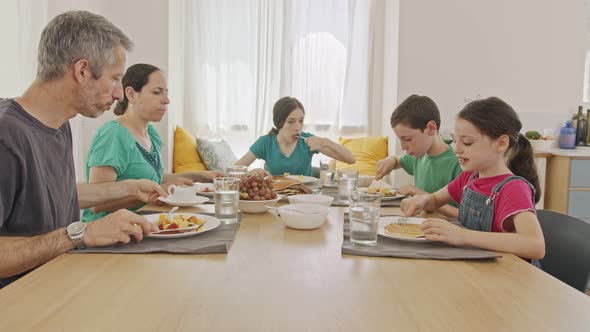 Family of five sitting at the table eating pancakes and fruits for breakfast alt
