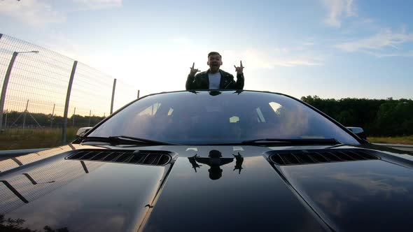 Handsome Funny Man Standing Out of Car Sunroof and Showing Crazy Emotions alt
