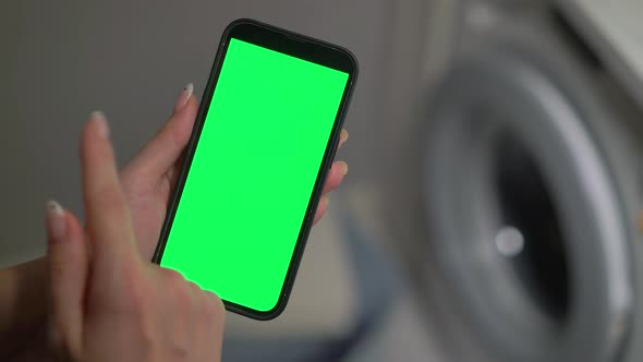 Woman Holds a Mobile Phone with a Green Chromakey Screen in Her Hands in Laundry