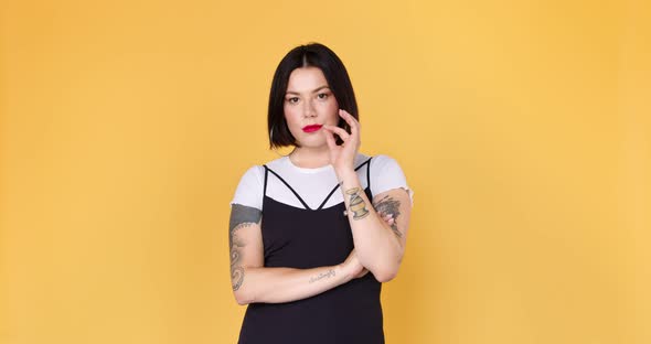 Smiling hipster woman with tattoo keep secret and looking at the camera on yellow background.  alt