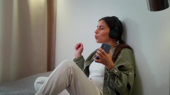 A Young Girl Listens to Music on Headphones at Home While Sitting on the Bed and Dances