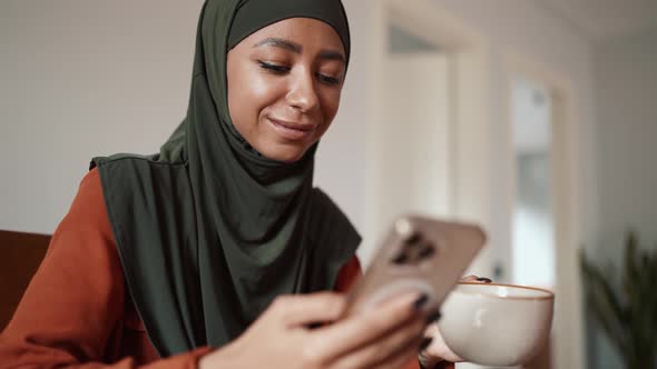 Young Muslim woman drinking tea and typing by phone alt
