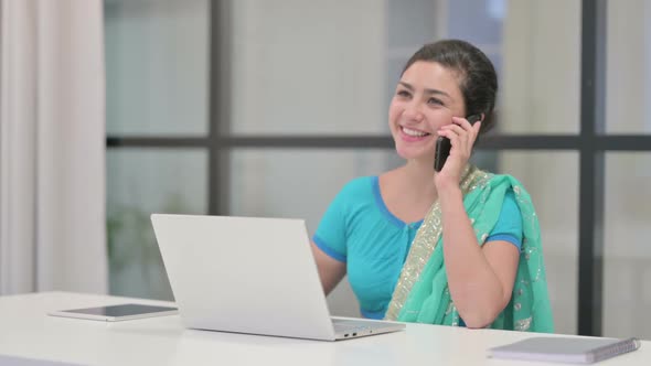 Indian Woman Talking on Phone While Using Laptop in Office alt