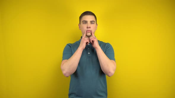 A Young Man Makes a Sad Smile on Her Face with His Fingers on a Yellow Background. Man in a Green alt