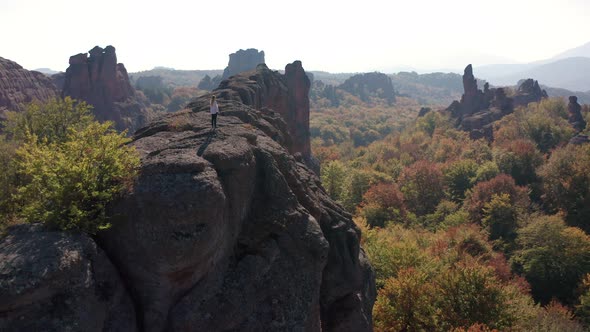 Aerial fly around a young woman standing at the adge of stunning rock formation alt