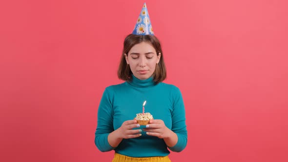 Young Woman in Birthday Hat Blowing Out Candle on Cake Isolated on Red Background alt