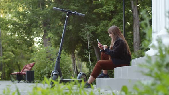 Woman is Sitting on Steps Park Next to an Electric Scooter and Typing Something in Her Phone Side alt
