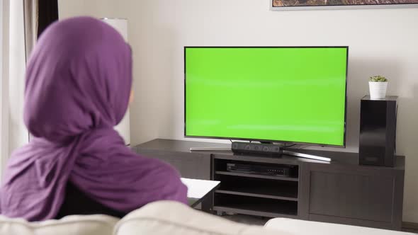 A Muslim Woman Watches TV with Green Screen and Skips Through Channels with a Remote Control alt