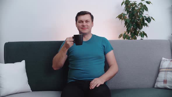 The man smiles happily, drinks coffee, sitting on the couch.