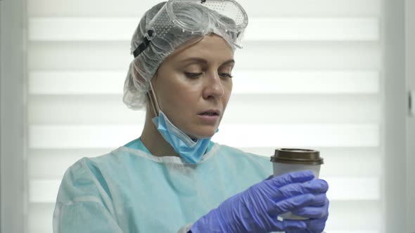 Tired Female Doctor or Nurse in a Protective Suit Drinks Coffee