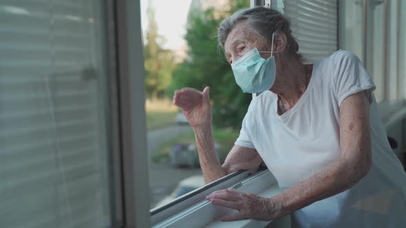 Lonely Senior Woman in Mask Stands By Window and Waves Her Hand to Family Who Came Visit Her at alt