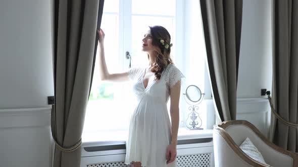 Young pregnant woman in white dress stand next to window in stylish room