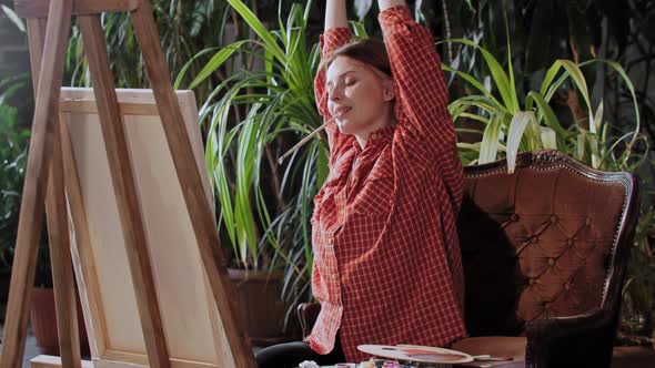 Artist in an Art Studio with Plants Young Pretty Woman Stretching Up and Starts Drawing a Painting alt