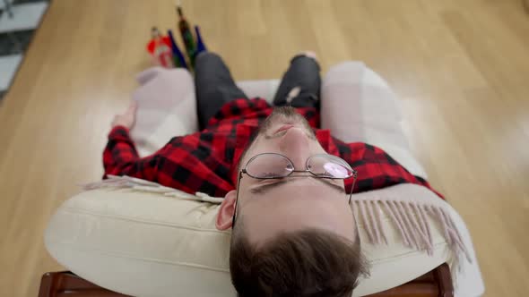 Top View of Drunken Young Man in Eyeglasses Leaning on Armchair with Blurred Alcohol Bottles alt