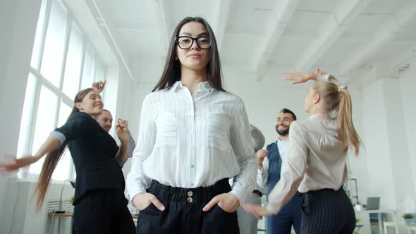 Young Asian Businesswoman with Serious Face Standing in Office Where People Men and Women Dancing alt