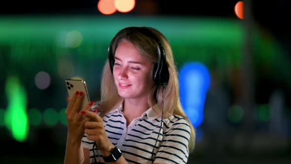 Cute Young Girl Chooses and Listens to Music Standing on the Street in the Night City