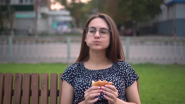 A Young Pregnant Girl is Eating a Burger While Sitting on a Bench in the Park alt