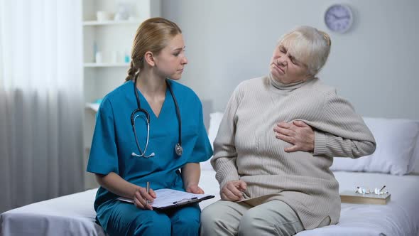 Young Doctor Examining Elderly Lady Complaining on Heart Pain, Writing Diagnosis alt