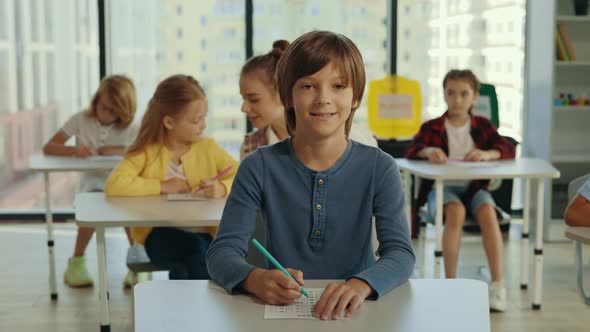 Portrait of the Male Smiled Pupil Looking at the Camera Sitting on the Desk and Writing His Exam on alt