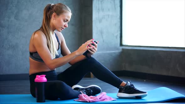 Woman in the Gym with Smartphone. She Sitting on the Mat and Using Smartphone alt