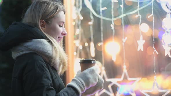 Young happy girl drinks hot coffee looks into the glowing shop window.