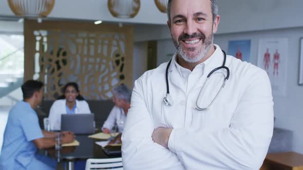 Portrait of caucasian male doctor smiling, with colleagues in discussion in background alt