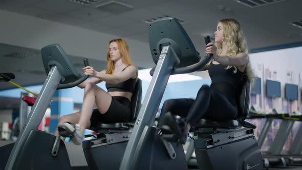 Gorgeous Blond Fit Young Woman Talking with Redhead Friend Training on Exercise Bikes alt