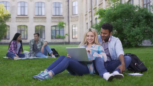 Multi-Ethnic Students Sitting on Grass and Watching Funny Video on Laptop alt