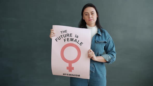 Slow Motion Portrait of Adult Woman Holding Feminist Sign Standing Against Grey Background alt