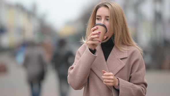 young girl in a coat standing on street and drinking hot drink