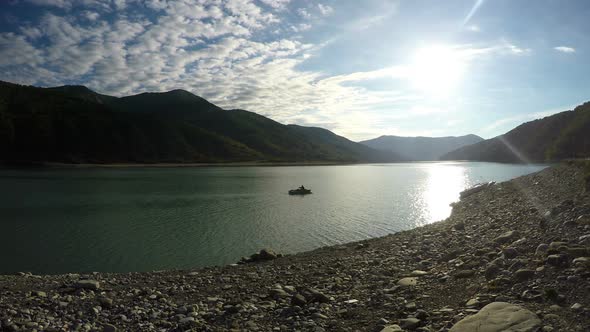 Man in Boat Rowing Down River With Mountains Around, Sunshine Reflected in Water alt