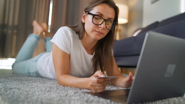Woman with Glasses Is Lying on the Floor and Makes an Online Purchase Using a Credit Card and Laptop alt