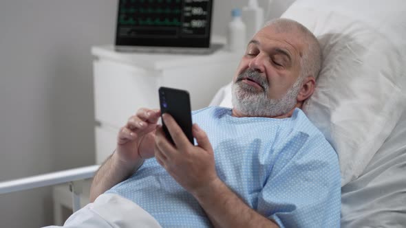 Hospital Ward Senior Man Resting in Bed Uses Smartphone for Video Call Conference Talk with Family alt