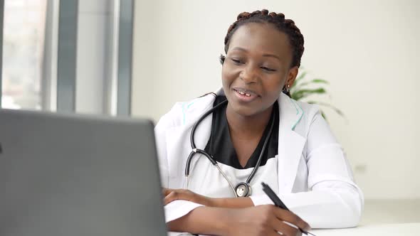 Young African American Woman Doctor Having Chat or Consultation on Laptop and Makes Some Notes alt