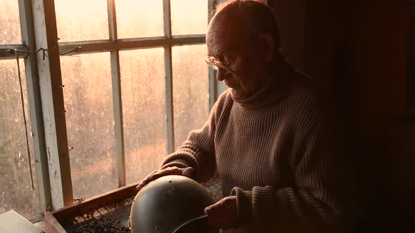 Elderly Man Glasses Picks Up Military Helmet Brushing Dust Off in House Window alt