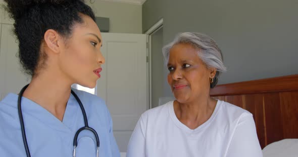 Front view of young mixed-race female doctor and senior black woman sitting on bed at home 4k alt