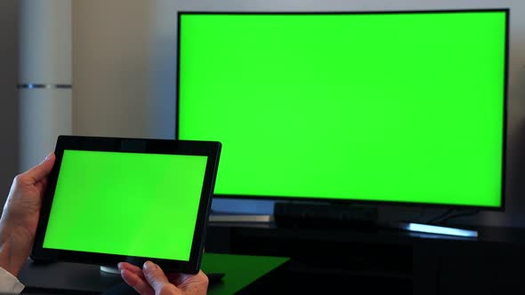Old Caucasian Woman Watches Television in Living Room and Holds Tablet in Hand alt