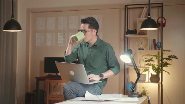 Designer Sits On His Desk Holds Laptop On The Knees Drinks Coffee While Working On The Project alt