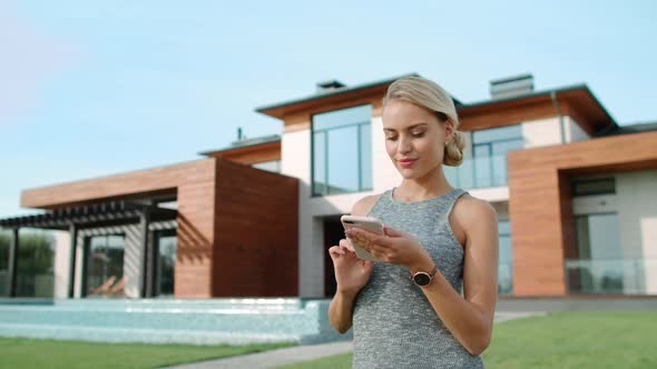Beautiful Woman Typing Mobile Near Luxury House Exterior. Successful Lifestyle alt