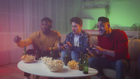 Cheerful Young Diverse Male Friends Playing Video Games with Joysticks Resting at Home in Evening alt