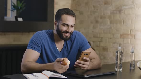 Handsome Young Middle Eastern Man Surfing Internet on Smartphone and Eating Apple. Portrait of Happy alt