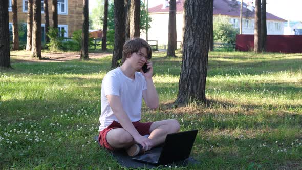 Young Man is Sitting on the Grass in Park with His Laptop and Talking on the Phone