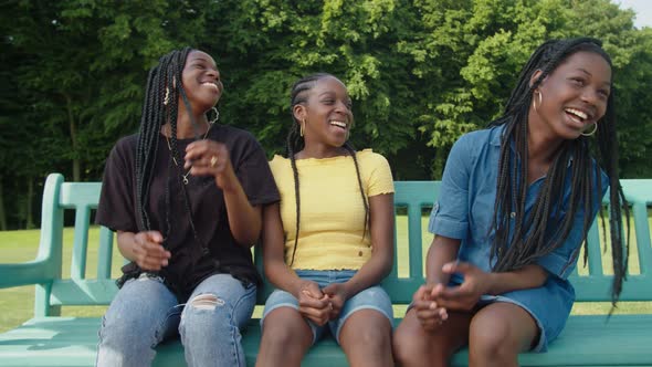Cheerful Adorable African Teenage Sisters Resting and Talking on Park Bench alt