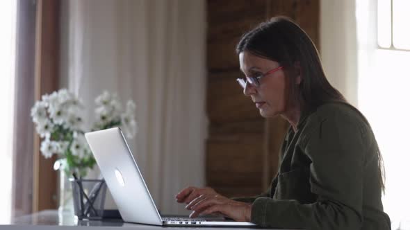 European Elderly Woman is Using Laptop for Remote Work Sitting at Table in Home Interior Spbi alt
