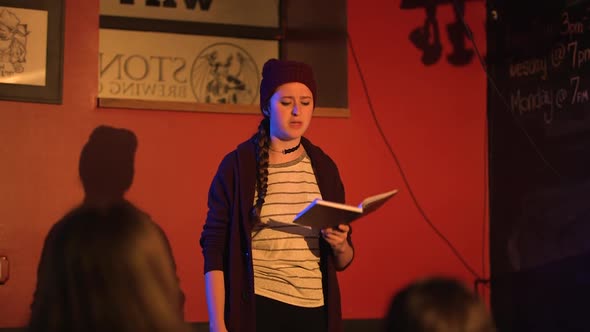Young women reads slam poetry book and walks off stage wearing red beanie alt