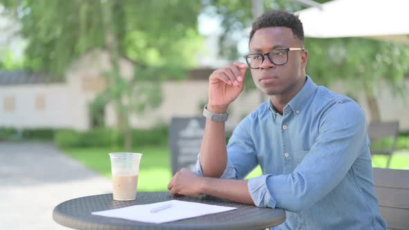 Pensive African Man with Coffee and Documents Thinking in Outdoor Cafe alt