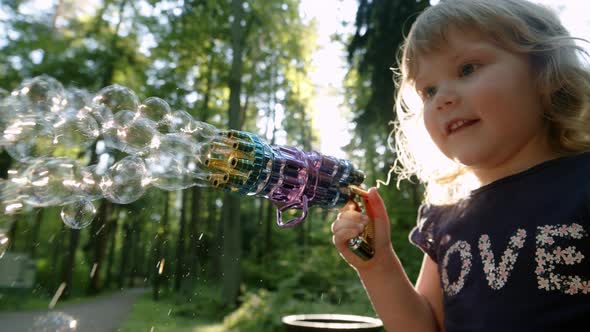 Happy Kid Blows Bubbles From Special Manual Bubble Machine Outdoors in Park alt
